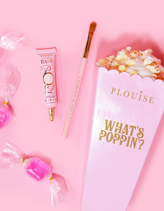A makeup brush and a tube of P.Louise Base next to a popcorn bucket surrounded by candies on a pink background.