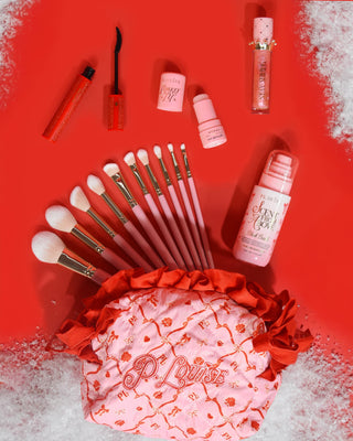 粉色圣诞 P.Louise 化妆包，上面有红色节日设计，顶部有化妆产品，包括化妆刷、唇油、睫毛膏等，红色背景有雪