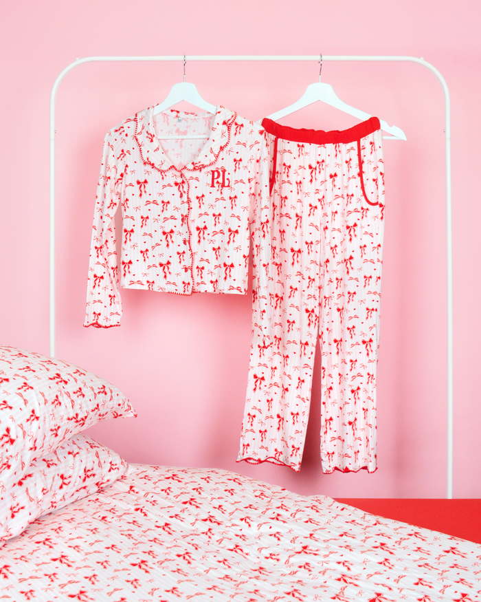 Girls P.Louise Christmas bow pyjama trousers set featuring an all over red bow design against pink and white stripes hanging on coat hangers in a pink bedroom with matching bedding