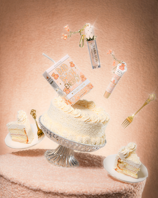 泰迪布桌上的蛋糕，椰子蛋糕上有 P.Louise Teddy Glitter Bad Bitch Energy Lip Duo，蛋糕周围漂浮着润唇膏和唇油 