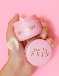 A person holding a pink jar of P.Louise Give It A Rest eye cream with the lid off, showing the product name and branding on the jar.