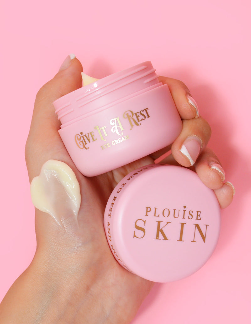 A person holding a pink jar of P.Louise Give It A Rest eye cream with the lid off, showing the product name and branding on the jar.